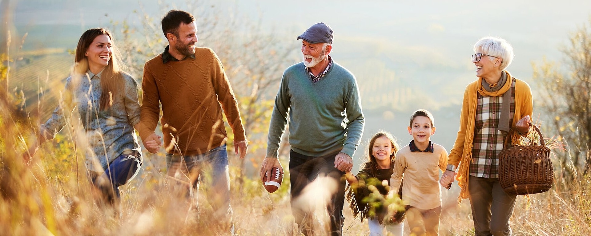 Eine sechsköpfige Familie, darunter zwei Kinder, geht gemeinsam durch ein malerisches, sonnenbeschienenes Feld. Sie tragen Herbstkleidung und lächeln, im Hintergrund sind sanfte Hügel zu sehen. Eine ältere Frau trägt einen Weidenkorb.