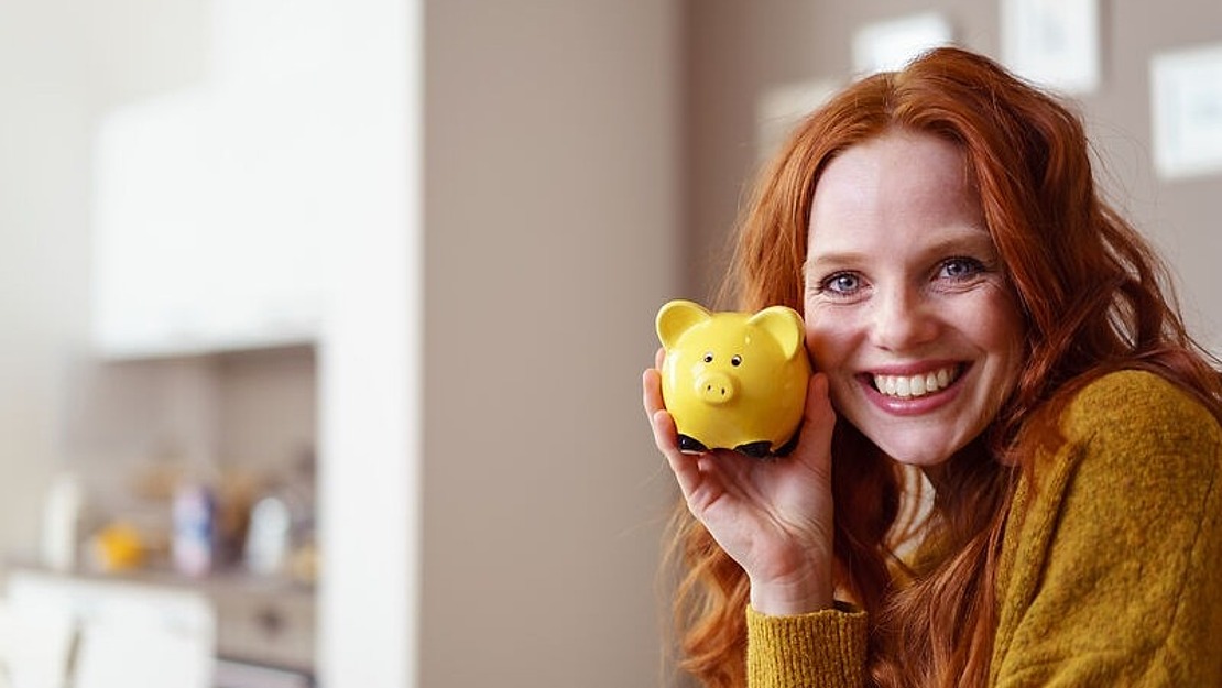 Eine lächelnde Frau mit langen roten Haaren, die einen senffarbenen Pullover trägt, hält ein gelbes Sparschwein vor ihr Gesicht. Der Hintergrund ist ein helles Kücheninterieur.