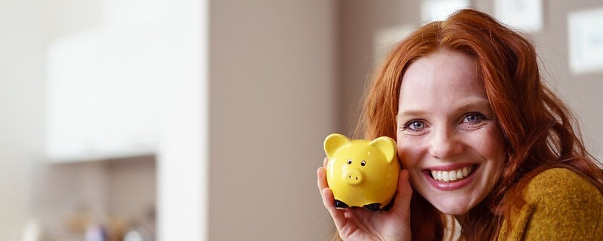 Eine lächelnde Frau mit langen roten Haaren, die einen senffarbenen Pullover trägt, hält ein gelbes Sparschwein vor ihr Gesicht. Der Hintergrund ist ein helles Kücheninterieur.