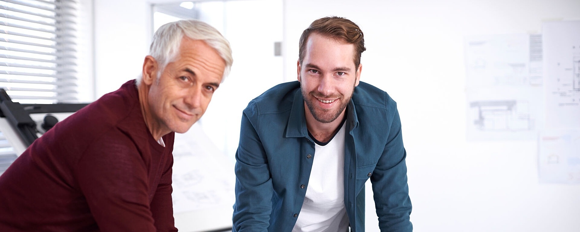 Zwei Männer beugen sich über einen Tisch in einem hellen Büroraum. Einer hat graue Haare und trägt einen kastanienbraunen Pullover, der andere hat braune Haare und trägt eine blaue Jacke. Sie lächeln beide, auf dem Tisch liegen Dokumente und ein Smartphone.