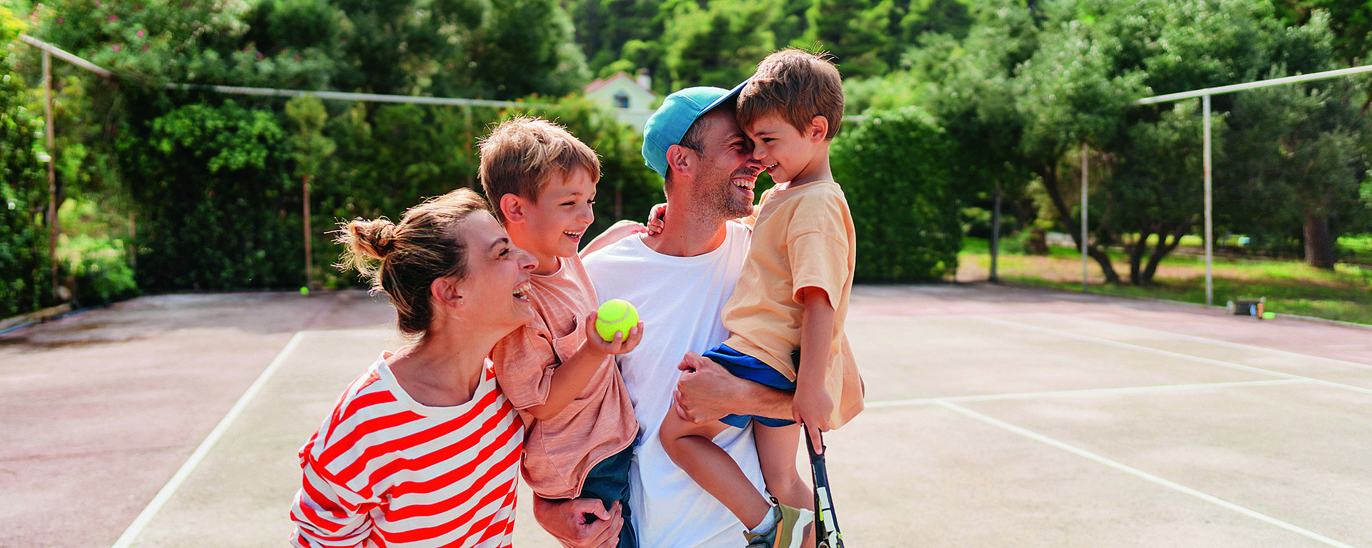 Eine fröhliche Familie auf einem Tennisplatz. Ein Erwachsener in einem gestreiften Hemd lächelt und hält ein Kind im Arm. Ein anderer Erwachsener hält einen Schläger und ein Kind im Arm. Im Hintergrund umgeben Bäume den Platz.