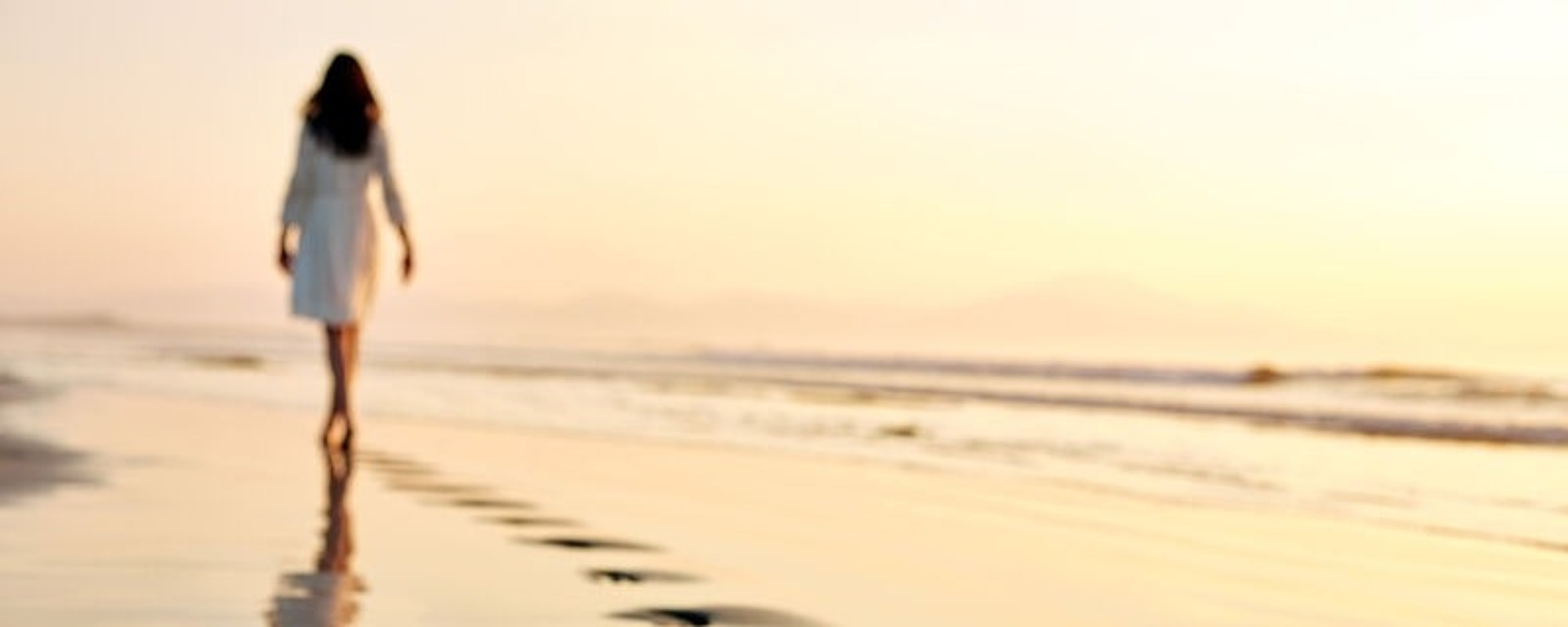 Eine Person in einem weißen Kleid geht an einem Sandstrand entlang und hinterlässt Fußspuren. Der Sonnenuntergang wirft einen warmen Schimmer auf den nassen Sand und schafft eine ruhige Atmosphäre. Am Horizont sind schwach Hügel in der Ferne zu erkennen.