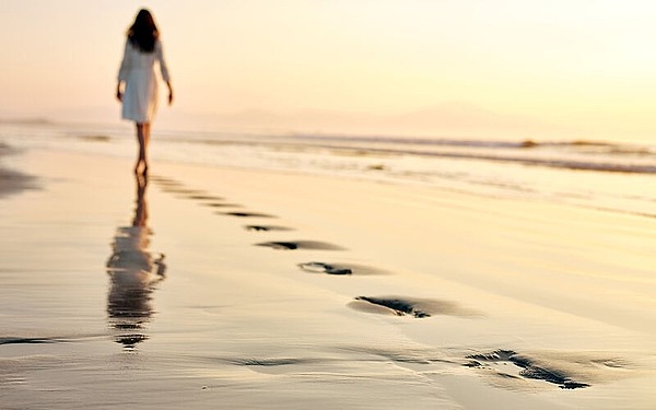 Eine Person in einem weißen Kleid geht an einem Sandstrand entlang und hinterlässt Fußspuren. Der Sonnenuntergang wirft einen warmen Schimmer auf den nassen Sand und schafft eine ruhige Atmosphäre. Am Horizont sind schwach Hügel in der Ferne zu erkennen.