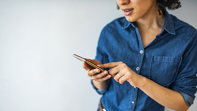 Eine Person, die ein blaues Jeanshemd trägt, benutzt ein Smartphone, hält es in einer Hand und berührt mit der anderen den Bildschirm vor einem schlichten hellen Hintergrund. Das Gesicht der Person ist teilweise sichtbar.