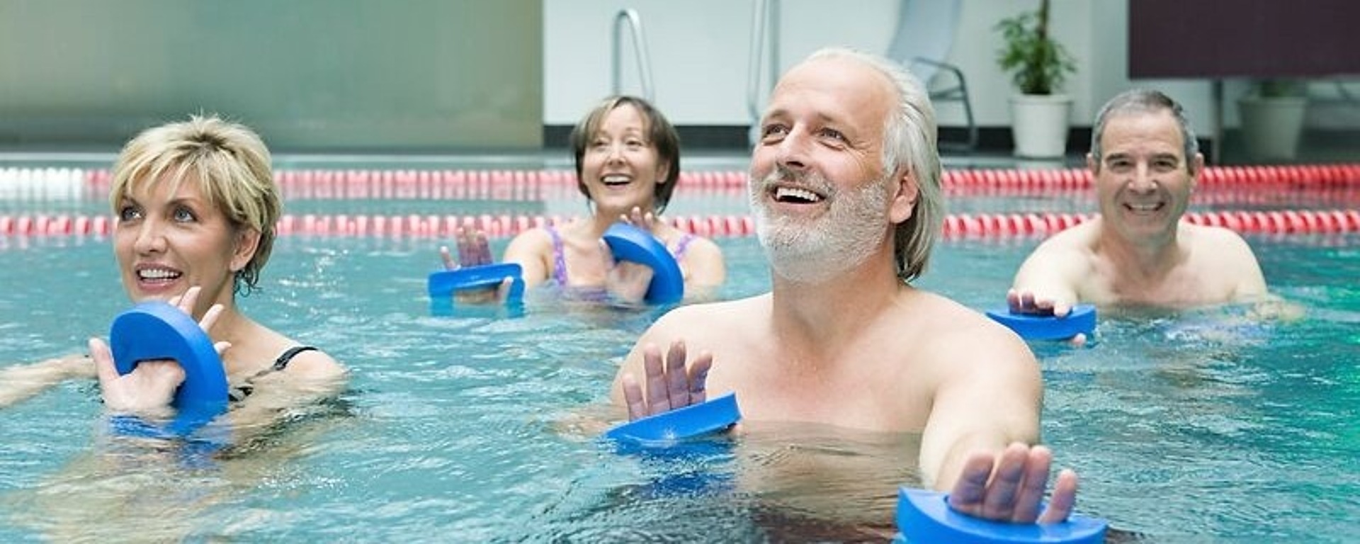 Vier lächelnde Menschen in einem Schwimmbecken machen Wassergymnastik. Jeder hält blaue Schaumstoffgewichte in der Hand. Sie wirken voller Energie und stehen in einer Reihe neben einer rot-weißen Bahnmarkierung. Im Hintergrund sind ein Liegestuhl und eine Pflanze zu sehen.