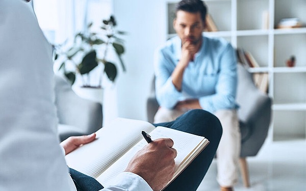 Eine Person sitzt mit einem Notizbuch und einem Stift da und macht sich möglicherweise Notizen, während eine andere Person mit nachdenklichem Gesichtsausdruck ihr gegenüber sitzt. Sie befinden sich in einem Raum mit Bücherregalen und einer Pflanze. Die Szene lässt auf ein Gespräch oder eine Therapiesitzung schließen.