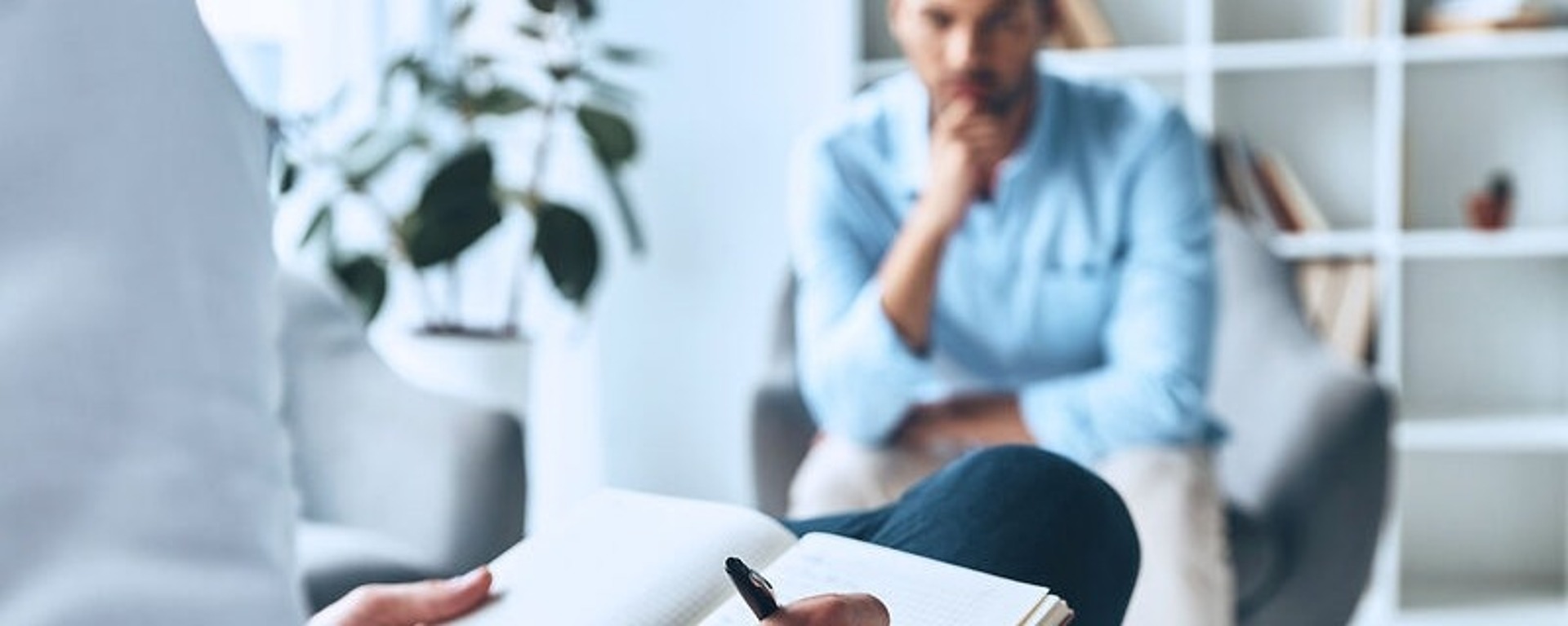 Eine Person sitzt mit einem Notizbuch und einem Stift da und macht sich möglicherweise Notizen, während eine andere Person mit nachdenklichem Gesichtsausdruck ihr gegenüber sitzt. Sie befinden sich in einem Raum mit Bücherregalen und einer Pflanze. Die Szene lässt auf ein Gespräch oder eine Therapiesitzung schließen.