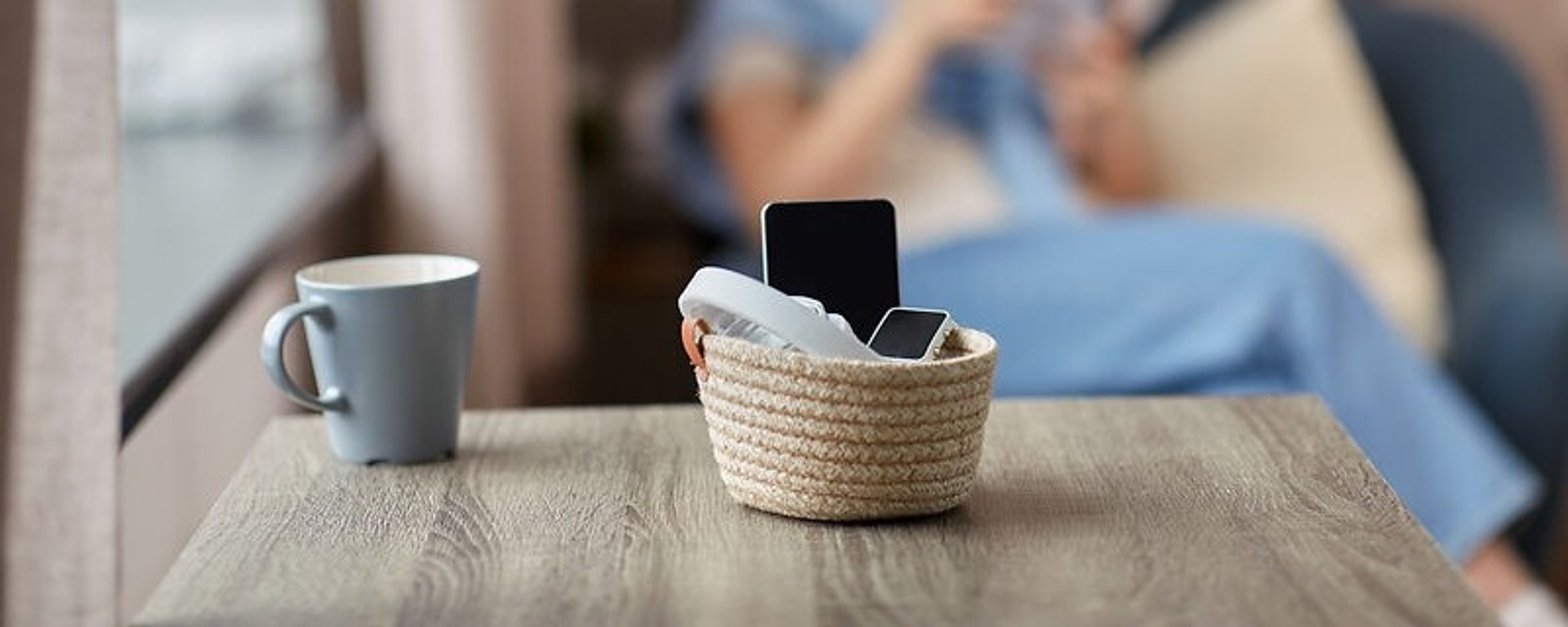 Ein Korb mit zwei Smartphones steht auf einem Holztisch. Daneben steht eine hellblaue Tasse. Im unscharfen Hintergrund sitzt eine Person in blauer Kleidung auf einem Sofa und liest ein Buch.