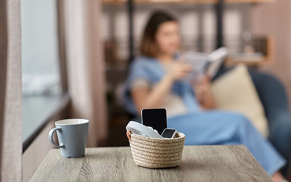 Ein Korb mit zwei Smartphones steht auf einem Holztisch. Daneben steht eine hellblaue Tasse. Im unscharfen Hintergrund sitzt eine Person in blauer Kleidung auf einem Sofa und liest ein Buch.