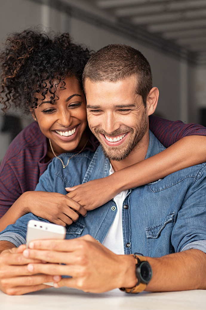 Ein lächelndes Paar sitzt an einem Tisch und schaut auf ein Smartphone. Die Frau hat lockiges Haar und umarmt den Mann von hinten. Der Mann, der ein Jeanshemd und eine Uhr trägt, hält das Telefon, während beide von dem, was sie sehen, entzückt zu sein scheinen.