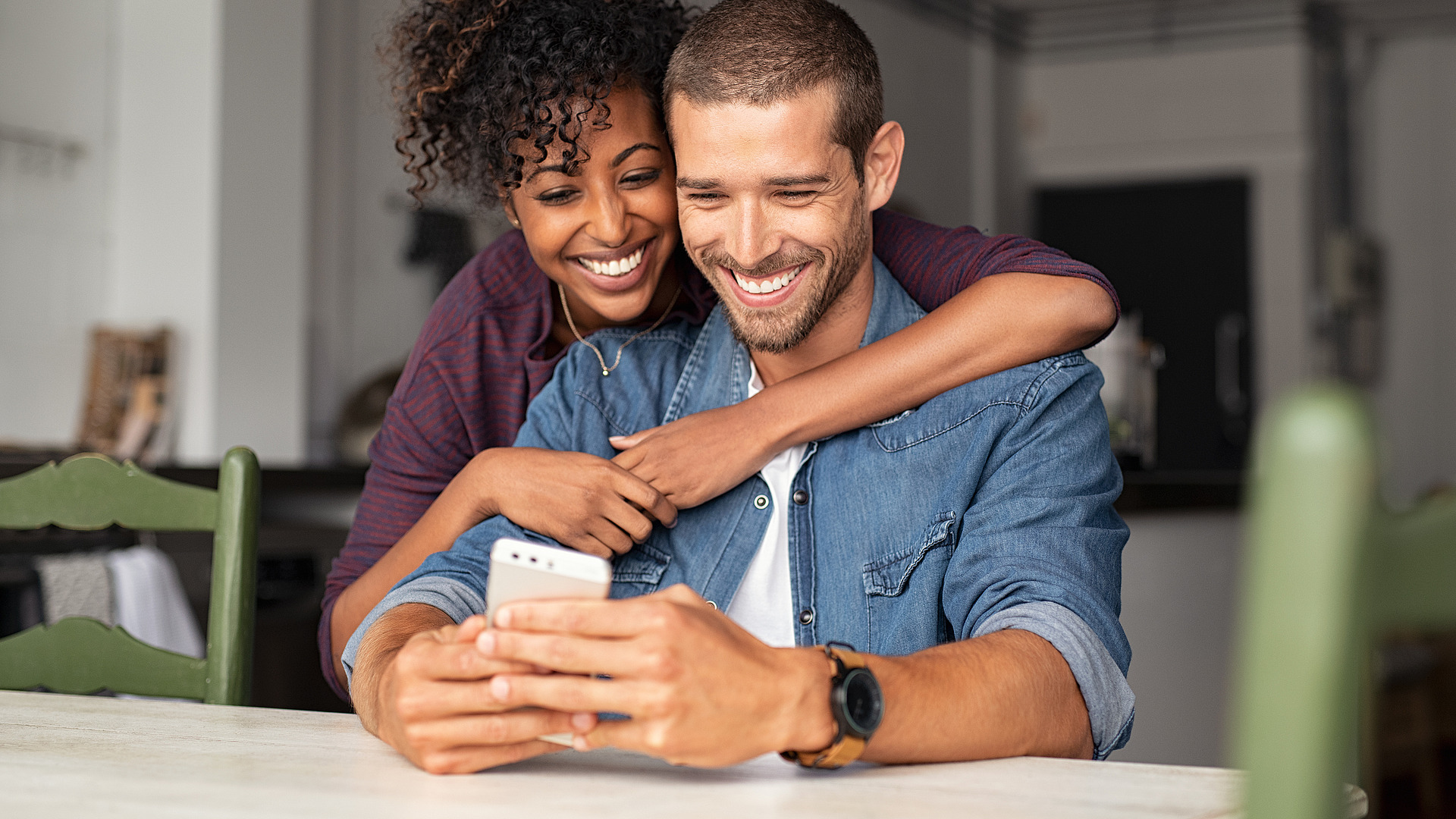 Ein lächelndes Paar sitzt an einem Tisch und schaut auf ein Smartphone. Die Frau hat lockiges Haar und umarmt den Mann von hinten. Der Mann, der ein Jeanshemd und eine Uhr trägt, hält das Telefon, während beide von dem, was sie sehen, entzückt zu sein scheinen.