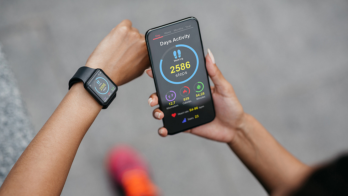 Person hält ein Smartphone, auf dem in einer Fitness-App 2.586 Schritte angezeigt werden, und trägt eine Smartwatch, auf der Zeit und Schritte angezeigt werden. Im Hintergrund ist ein unscharfer Schuh zu sehen.