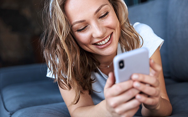 Lächelnde Frau, die auf einem grauen Sofa liegt und auf ihr Smartphone schaut.